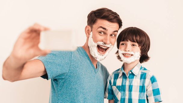 Young Father Teaches Little Son to Shave at Home. Foam on Face. Shaving Tool. Razor with Blade. Happy Family Concept. Young Boy in Shirt. Hygiene and Skincare Concepts. Taking Selfie. - Foto, afbeelding