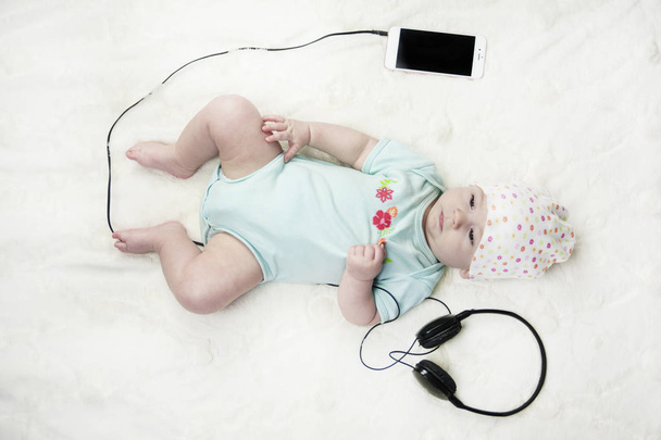 Bright little beautiful newborn baby lying on the bed listening to music and next to the headset with the phone close-up - Foto, imagen