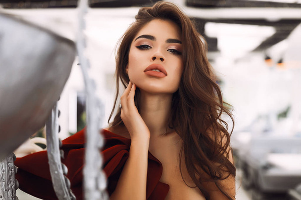 Amazing brunette model with beautiful makeup on the face, big lips and soft skin. Wearing burgundy dress with decollete and open shoulder. Stylish curly hair lying on the shoulder, hand touching neck - Photo, Image