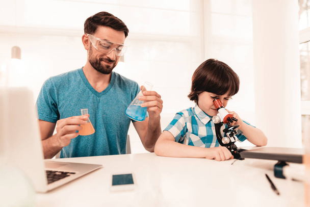Youn Bearded Father Teaching Son in Shirt at Home. Education at Home. Using Microscope. Studding Chemistry. White Table in Room. Sitting Boy. Young Father. Lesson at Home. Education Concept. - Foto, immagini