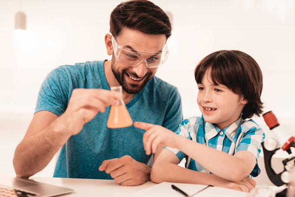 Youn Bearded Father Teaching Son in Shirt at Home. Education at Home. Using Microscope. Studding Chemistry. White Table in Room. Sitting Boy. Young Father. Lesson at Home. Education Concept. - 写真・画像