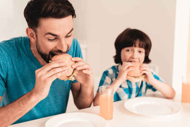 Smiling Son and Father Have Lunch in Kitchen. Boy in Shirt. Modern Kitchen. Sitting Boy. Boy with Spoon. Breakfast in Morning. White Table in Kitchen. Burger in Hands. Young Father. - Φωτογραφία, εικόνα