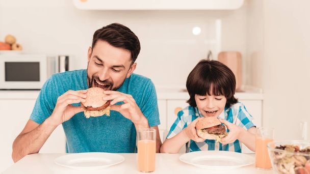 Smiling Son and Father Have Lunch in Kitchen. Boy in Shirt. Modern Kitchen. Sitting Boy. Boy with Spoon. Breakfast in Morning. White Table in Kitchen. Burger in Hands. Young Father. - Photo, Image