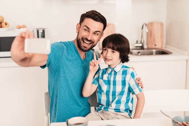 Happy Father and Son Using Smartphone at Home. Smiling Boy. Modern Kitchen. Sitting Boy. Boy in Shirt. Breakfast in Morning. White Table in Kitchen. Using Digital Device. Happy Family. - Foto, afbeelding