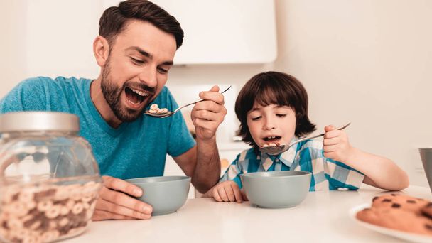 Smiling Son and Father Have Breakfast in Kitchen. Boy in Shirt. Modern Kitchen. Sitting Boy. Boy with Spoon. Breakfast in Morning. White Table in Kitchen. Gray Bowl on Table. Young Father. - Foto, Imagem