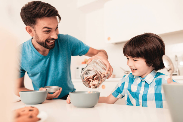 Smiling Son and Father Have Breakfast in Kitchen. Boy in Shirt. Modern Kitchen. Sitting Boy. Boy with Spoon. Breakfast in Morning. White Table in Kitchen. Gray Bowl on Table. Young Father. - Фото, изображение