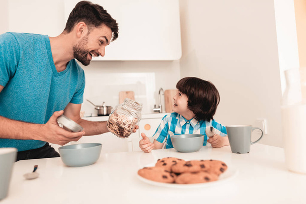 Smiling Son and Father Have Breakfast in Kitchen. Boy in Shirt. Modern Kitchen. Sitting Boy. Boy with Spoon. Breakfast in Morning. White Table in Kitchen. Gray Bowl on Table. Young Father. - Photo, Image
