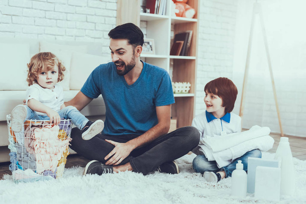 Father is Engaged in Raising Children. Father is Teaching a Sons a Cleaning. People Want to Wash a Dirty Clothes. Younger Son is Sitting in Basket with Clothes. People is Located in Bedroom on Carpet. - Φωτογραφία, εικόνα
