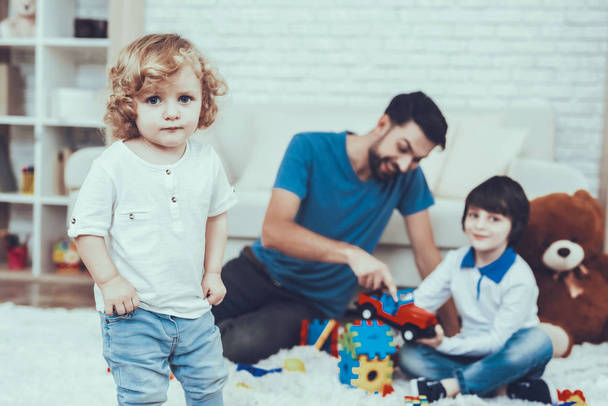 Man Spends Time with His Sons. Father is Engaged in Raising Children. Father and Eldest Sons is Playing with Toys on Carpet. Younger Son is Standing Alone. People is Located in Bedroom. - Φωτογραφία, εικόνα