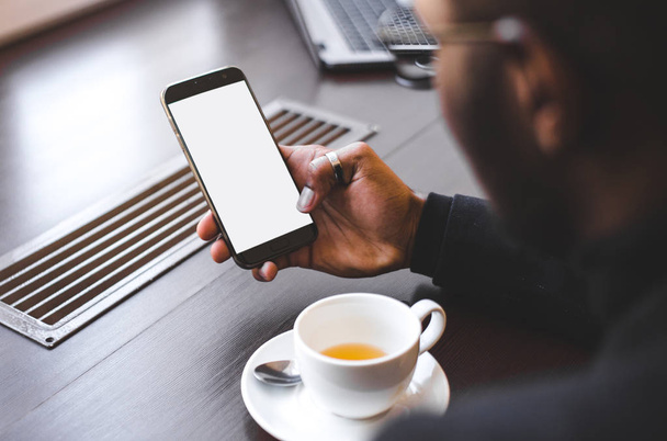 Un Afro-Américain assis dans un café, buvant du café et travaillant derrière un téléphone portable. Maquette écran
 - Photo, image