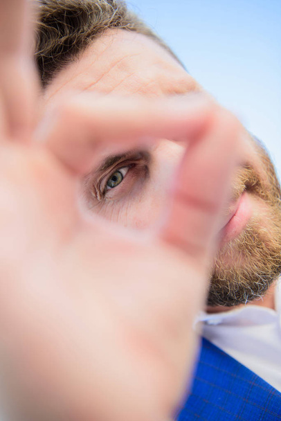 Guy looks suspicious through ok gesture close up. Excellent concept. Spying watching and observing. Man bearded businessman formal suit watching through fingers. I am watching you. Perfect gesture - Photo, image