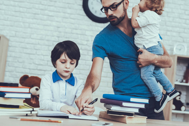 Father of Boys is Engaged in Raising Children. Father is Doing a Homework with Eldest Son. Man is Holding a Younger Son in Arm. Eldest Son is Sitting at Table. People Located at Home. - 写真・画像