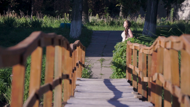 junge Frau mit weißem Fächer im weißen Kleid geht an einem sonnigen heißen Tag auf die Holzbrücke - Filmmaterial, Video