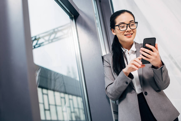Lächelnde Geschäftsfrau steht am Fenster und berührt Smartphone-Bildschirm - Foto, Bild