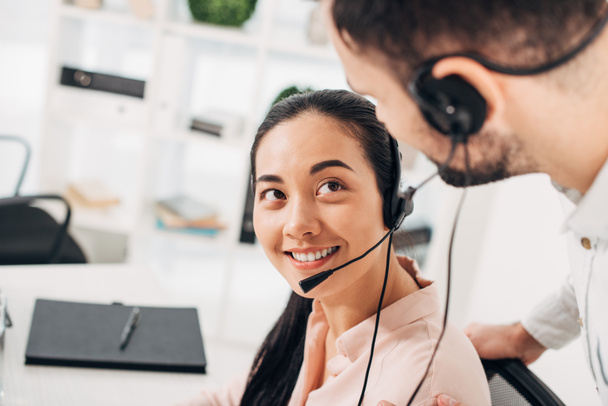 attractive call center operator smiling and looking at colleague in office - Photo, Image