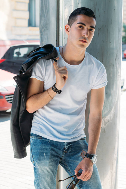 νεαρό, όμορφος άντρας σε ένα λευκό T-shirt, μαύρο, δερμάτινο σακάκι και μπλε, σκισμένα τζιν είναι στο δρόμο, μοντέλο, πόλη με τα πόδια, ζωντανή style.in Λευκά αθλητικά παπούτσια. με ένα ρολόι στο χέρι, λαμπερός ήλιος λάμπει, καλοκαίρι - Φωτογραφία, εικόνα