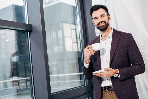 handsome businessman standing by window and holding coffee cup - Фото, изображение