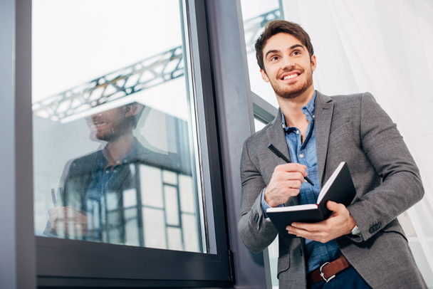 χαμογελώντας επιχειρηματίας στέκεται δίπλα σε παράθυρο και κρατώντας Ανοίξτε το σημειωματάριο στο γραφείο - Φωτογραφία, εικόνα