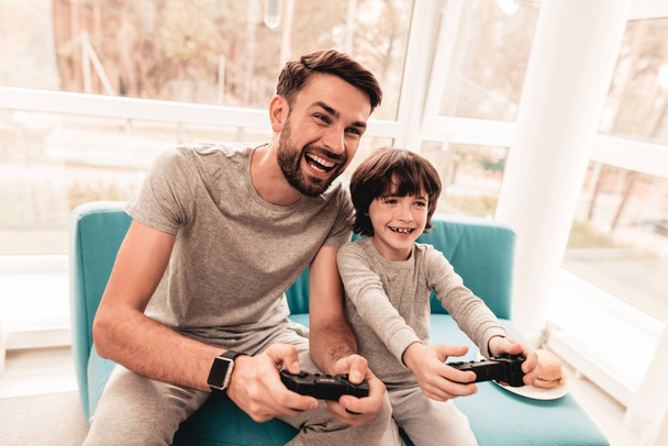 Father and Son Sitting and Playing on Console. Sitting Boy. Young Father. Playing on Console. Joystick in Hands. Young Bearded Man at Home. Sitting on Blue Sofa. Happy Family Concept. - Foto, Bild