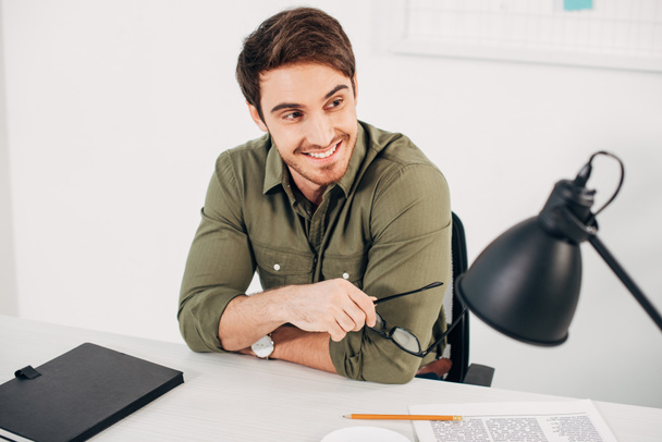 guapo gerente de oficina sentado en el escritorio, sonriendo y sosteniendo gafas
 - Foto, Imagen