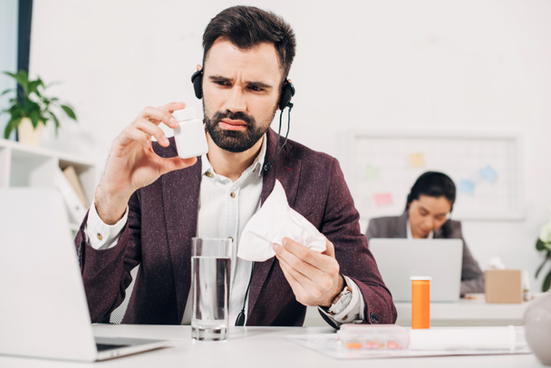 operador de call center doente sentado na mesa e segurando garrafa pílula e guardanapo no escritório
 - Foto, Imagem