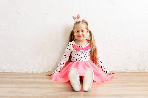 Portrait of cute little five year old girl with long blond hair wearing easter bunny ears on head, colorful polka dot shirt and pink tutu skirt. White wall backgroung, copy space, close up. - Photo, Image