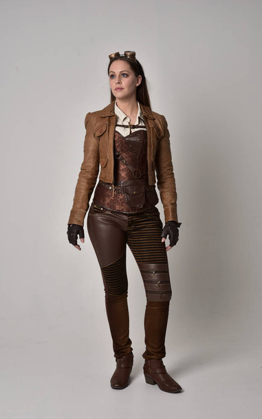 Portret van de volledige lengte van brunette meisje bruinleren steampunk outfit dragen. staande pose op de grijze studio achtergrond. - Foto, afbeelding