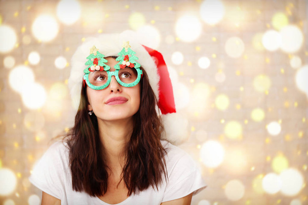 Beautiful young woman wearing santa claus furry hat & masquerade glasses for Chritmas celebration event. Attractive female being silly, making faces at dress up party. Background, close up, copy space - Foto, imagen