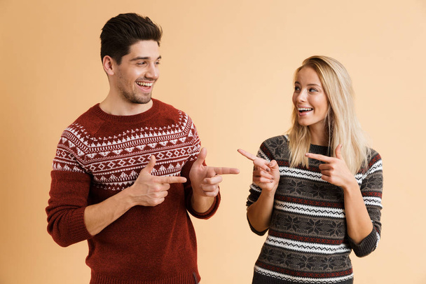 Retrato de um jovem casal feliz vestido com suéteres de pé juntos isolados sobre fundo bege, apontando um para o outro
 - Foto, Imagem