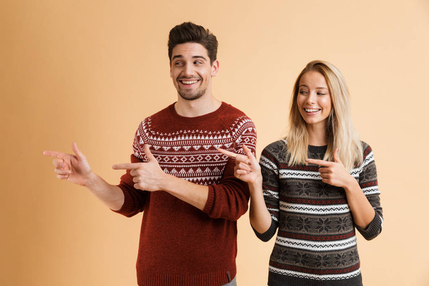 Portrait of a happy young couple dressed in sweaters standing together isolated over beige background, pointing away - Foto, afbeelding