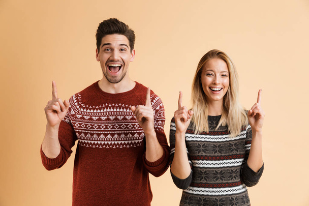 Portrait of a happy young couple dressed in sweaters standing together isolated over beige background, pointing up - Foto, imagen