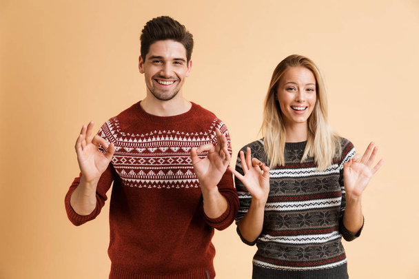 Portrait of a happy young couple dressed in sweaters standing together isolated over beige background, showing ok gesture - Foto, Imagem