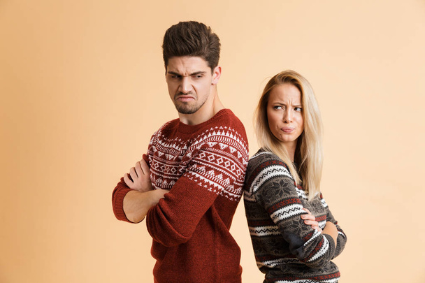 Portrait of an angry young couple dressed in sweaters standing together isolated over beige background, arms folded - Φωτογραφία, εικόνα