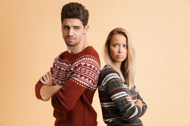 Πορτρέτο του ένα απογοητευμένος νεαρό ζευγάρι που ντυμένος με πουλόβερ, στέκεται μαζί απομονωμένη μπεζ φόντο, χέρια διπλωμένα - Φωτογραφία, εικόνα