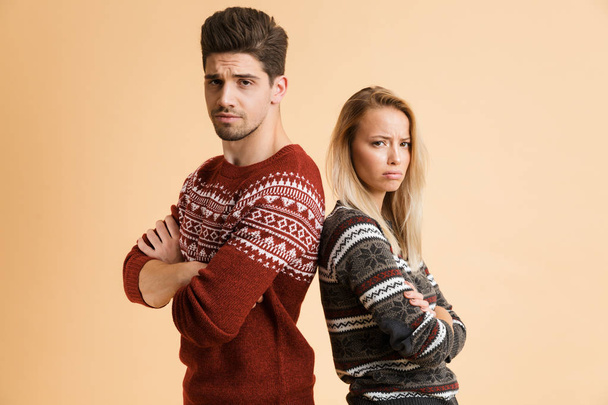 Portrait of an upset young couple dressed in sweaters standing together isolated over beige background, arms folded - Φωτογραφία, εικόνα
