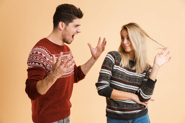 Portrait of a young couple dressed in sweaters standing together isolated over beige background, having an argument - Φωτογραφία, εικόνα
