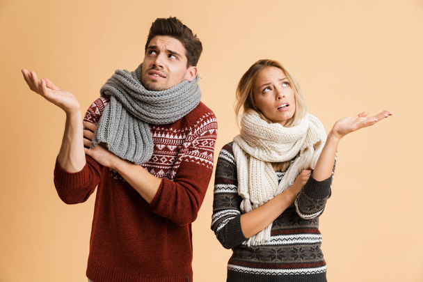 Portrait of a frozen young couple dressed in sweaters and scarves standing together isolated over beige background, shaking, outstretched hands - Фото, зображення