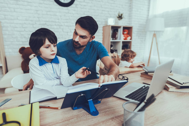 Father of Boys is Engaged in Raising Children. Father is Doing a Homework with Eldest Son. Younger Son is Playing on Background. People is Sitting at Table. People Located at Home. - Φωτογραφία, εικόνα