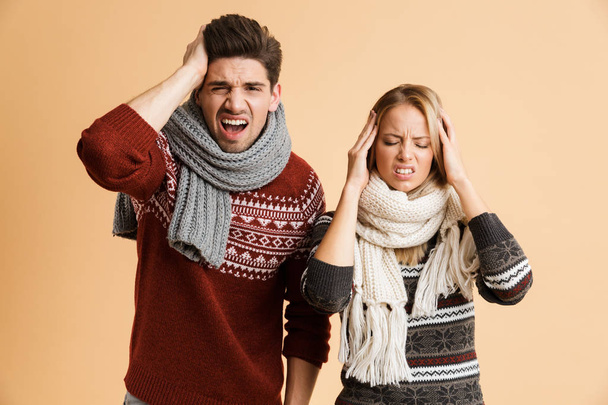 Portrait of a frozen young couple dressed in sweaters and scarves standing together isolated over beige background, having a headache - Fotografie, Obrázek