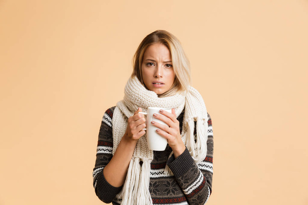 Portrait of an upset girl dressed in sweater and scarf isolated over beige background, holding cup of hot tea - Фото, изображение
