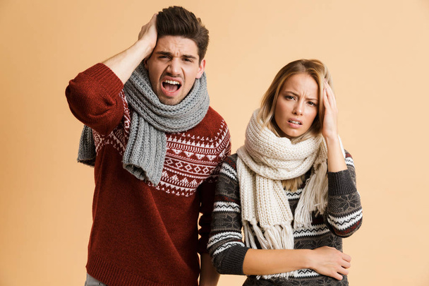 Portrait of a sick young couple dressed in sweaters and scarves standing together isolated over beige background, having a headache - Fotoğraf, Görsel