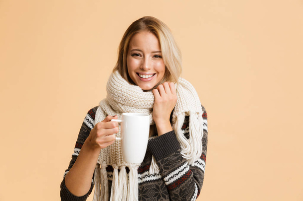 Portrait of a happy girl dressed in sweater and scarf isolated over beige background, holding cup of hot tea - Foto, immagini