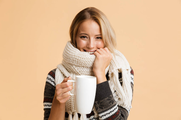Portrait of a smiling girl dressed in sweater and scarf isolated over beige background, holding cup of hot tea - Φωτογραφία, εικόνα