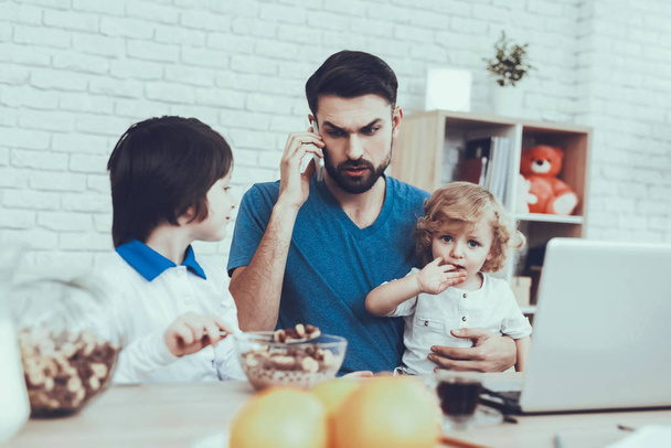 Father is Engaged in Raising Children. Eldest Son is Eating a Breakfast. Father is Working and Talking on Cellphone. Man is Holding a Younger Son. People is Sitting at Table. People Located at Home. - Фото, зображення