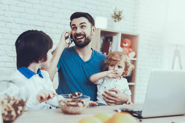 Father is Engaged in Raising Children. Eldest Son is Eating a Breakfast. Father is Working and Talking on Cellphone. Man is Holding a Younger Son. People is Sitting at Table. People Located at Home. - Foto, immagini