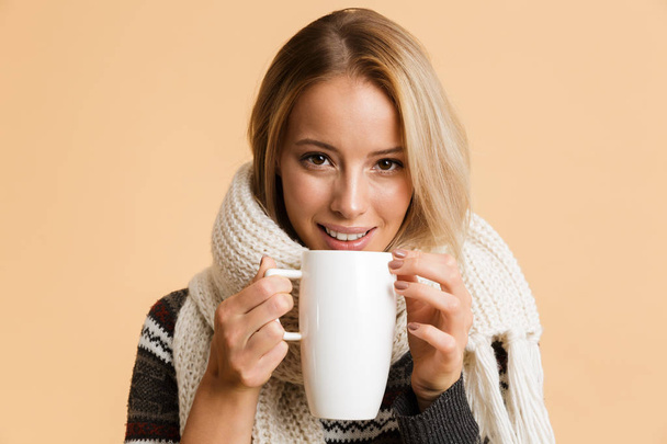 Close up portrait of a happy girl dressed in sweater and scarf isolated over beige background, holding cup of hot tea - Photo, Image