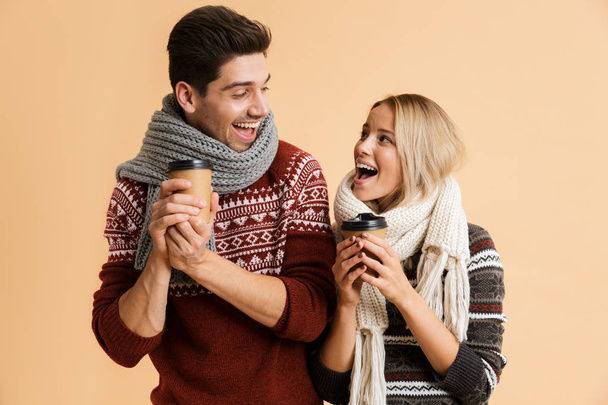 Portrait of a happy young couple dressed in sweaters and scarves standing isolated over beige backgorund, holding takeaway coffee cups - Foto, Imagem