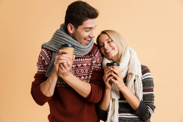 Portrait of a loving young couple dressed in sweaters and scarves standing isolated over beige backgorund, holding takeaway coffee cups - Foto, Imagem