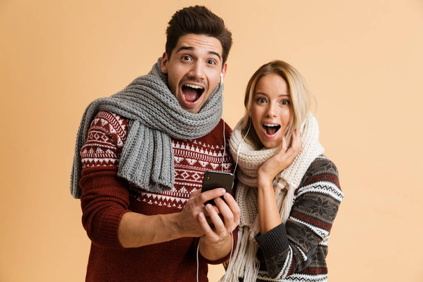 Image of shocked excited young loving couple in sweaters using mobile phone listening music isolated over beige background. - Valokuva, kuva