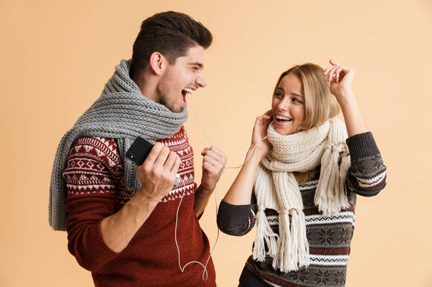 Portrait of a cheerful young couple dressed in sweaters and scarves standing isolated over beige backgorund, listening to music with earphones and dancing - Valokuva, kuva